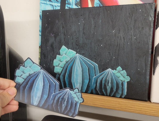 A hand holds a cutout featuring a geometric, cactus-like design in blue hues. In the background, the Crystal Succulent Weatherproof Decal/Sticker by InkBird Print Studio LLC decorates a canvas with similar motifs against a dark, starry sky and rests on a wooden shelf. The air-egress vinyl finish adds an intriguing touch to this captivating scene.