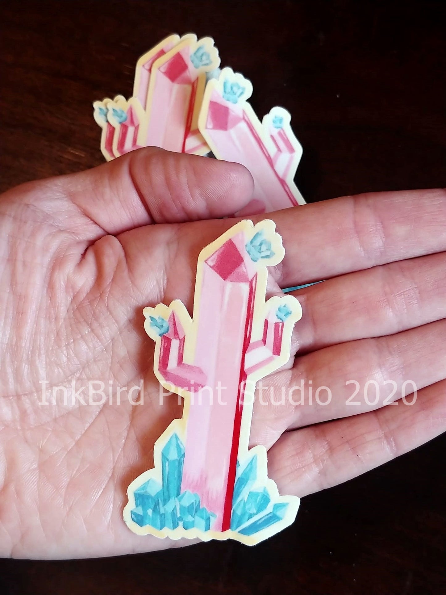 A hand displays a vibrant Crystal Cactus Decal/Sticker, featuring pink geometric crystals with blue accents set against a dark background. The brand name InkBird Print Studio LLC is visible on the hand. This 3"x 1.6" sticker is perfect for decorating laptops, gifts, water bottles, mirrors, lockers, and more!