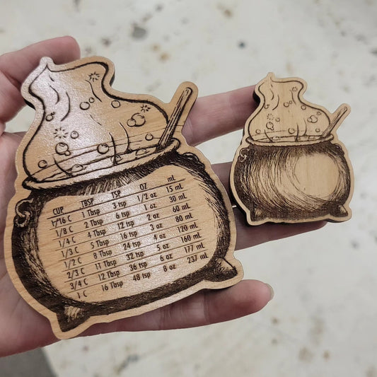 A photo showing two wood magnets. On the left is a large magnet 4" x 3.25" with kitchen conversions such as 1 cup is 16 tablespoons. On the right is a smaller version of the same cauldron without conversions.
