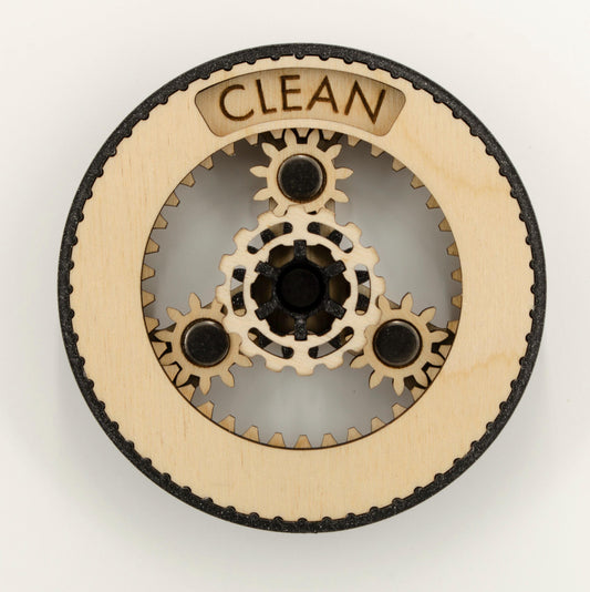 A round dishwasher magnet with rotating planetary gears made of laser-cut wood, enclosed in a 3D-printed PLA housing. The gears can be rotated to reveal either 'CLEAN' or 'DIRTY' in an engraved display window at the top, indicating the dishwasher's status.