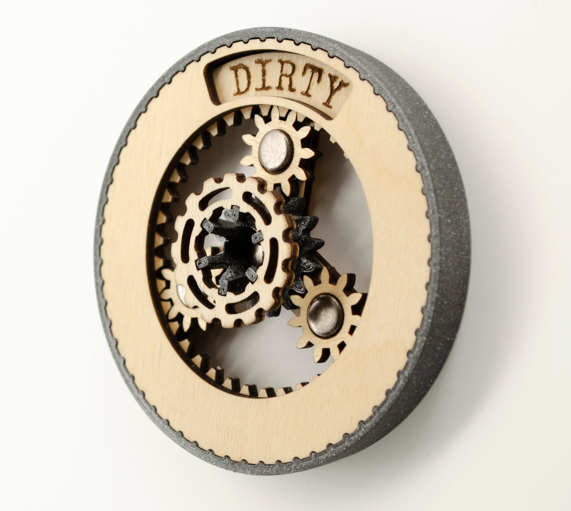 An angled view of a round dishwasher magnet with rotating planetary gears made of laser-cut wood, enclosed in a 3D-printed black PLA housing. The gears can be rotated to reveal either 'CLEAN' or 'DIRTY' in an engraved display window at the top, indicating the dishwasher's status.