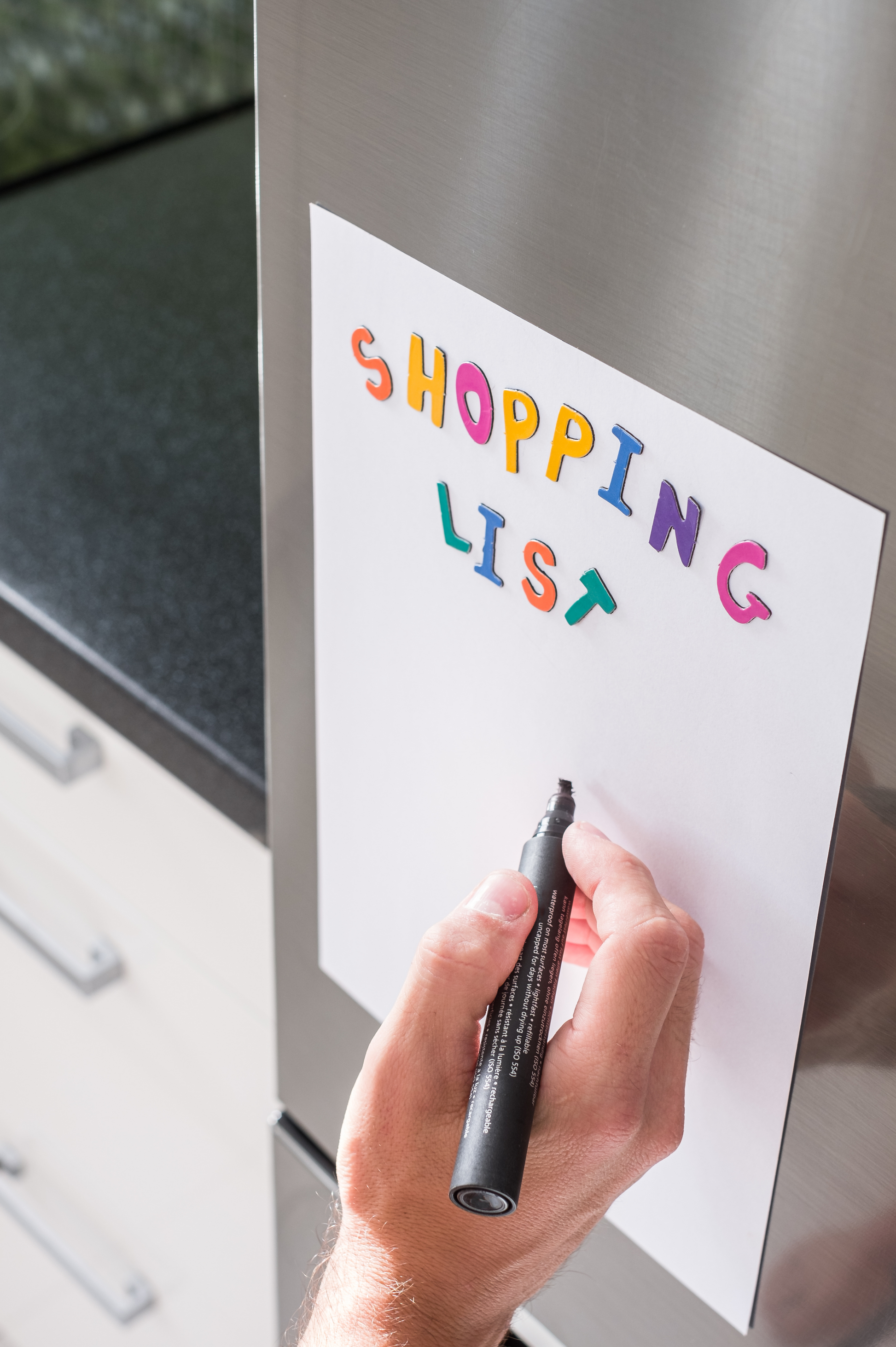 A hand holding a black marker writes on a white label titled "Shopping list' on a stainless steel refrigerator.