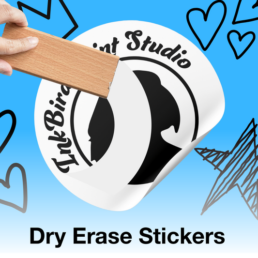 A hand holds a wooden block against a white circular sticker that reads 'InkBird Print Studio' with a black bird silhouette. The sticker is shown on a bright blue background with doodled heart and line drawings, and the text 'Dry Erase Stickers' appears at the bottom.