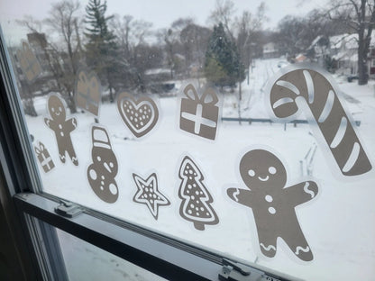 Image of The Holiday Cookie Static Cling Set by InkBird Print Studio LLC on a window showcasing their crisp printing. These are a renter-friendly way to decorate your window with gingerbread men, candy canes, a snowman, Christmas trees, hearts, and gift boxes. Outside of the window, you can enjoy the view of a snowy landscape adorned with trees and houses.