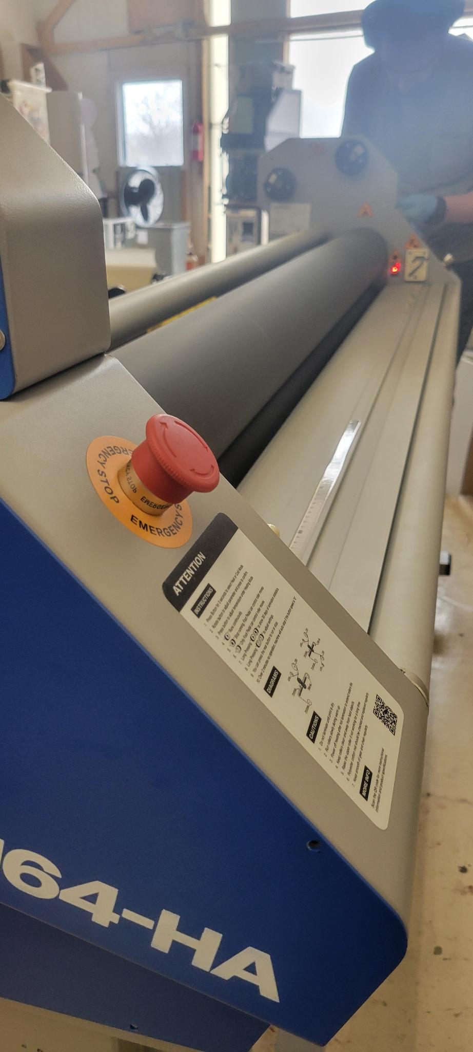 A brand new wide-format Ally Armour Laminator, 64 inches wide, photographed at an angle. A worker is in the background, adjusting a setting on the lamination machine.
