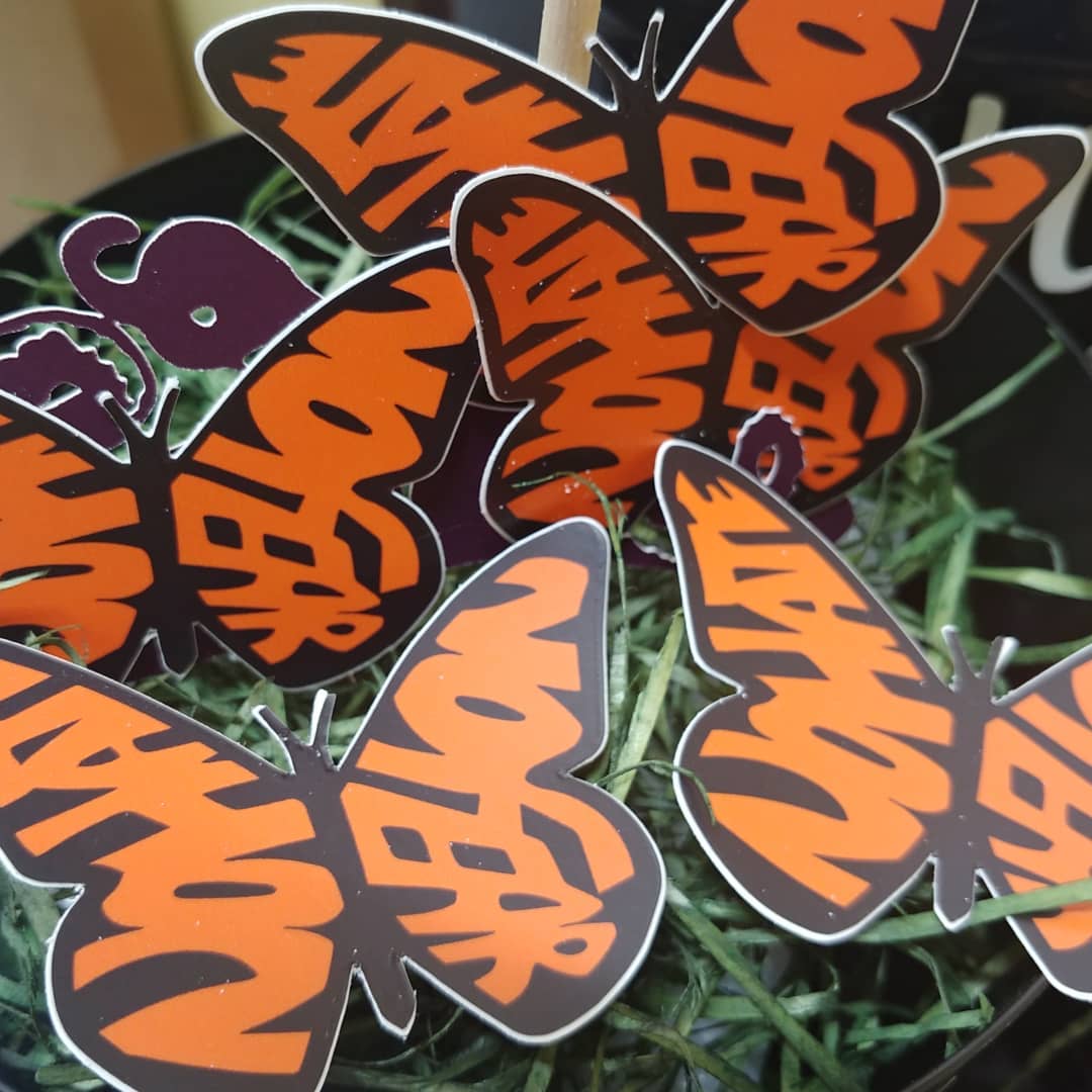 Scattered through in a pile of easter grass for a display: our No Hate No Fear Butterfly Decals. The wings, instead of the usual shapes seen on a Monarch Butterfly, spell out the words "No Hate" and "No Fear" in orange. The text is bold and stands out against the design, emphasizing a message of peace and positivity. This is a pro-immigration decal.