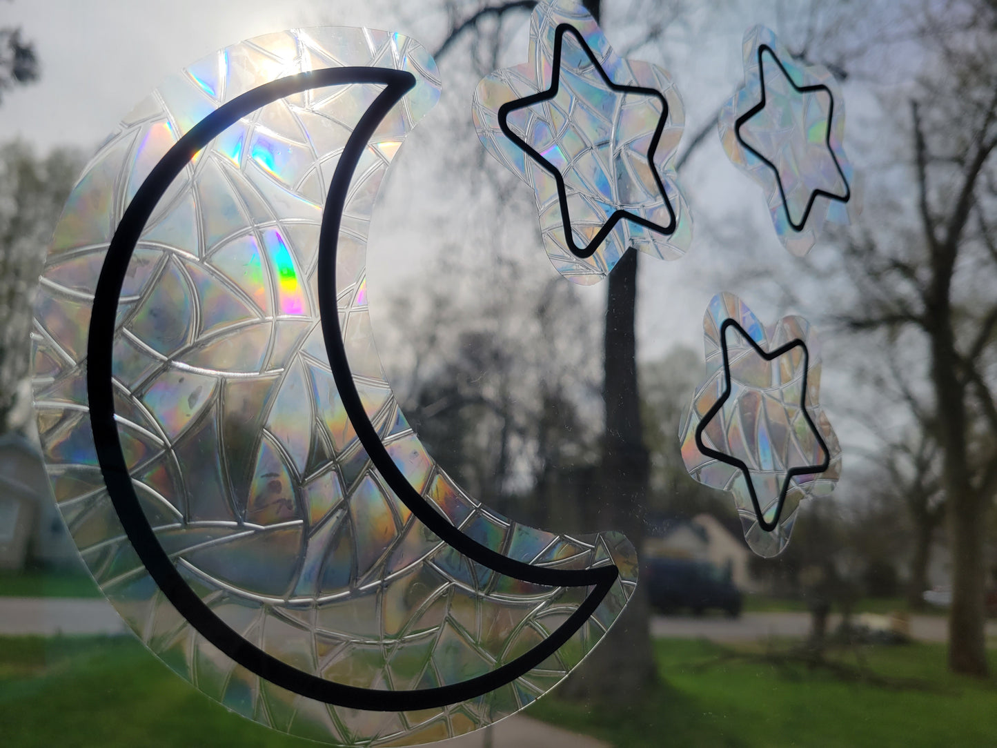 A close-up photo of the Moon and Stars Suncatcher Window Cling set by InkBird Print Studio. Black vinyl applied to a suncatcher style material, contour cut into moon and start shapes. Casting rainbows into the room as the sun hits it.
