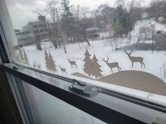 Image of the Winter Scene Static Cling Set by InkBird Print Studio LLC on a window showcasing their crisp printing and layer-ability. These are a renter-friendly way to decorate your window, featuring snowy hills, deer, pine trees, and a moon, all printed in white ink on clear static cling. Outside of the window, you can enjoy the view of a snowy landscape adorned with trees and houses.