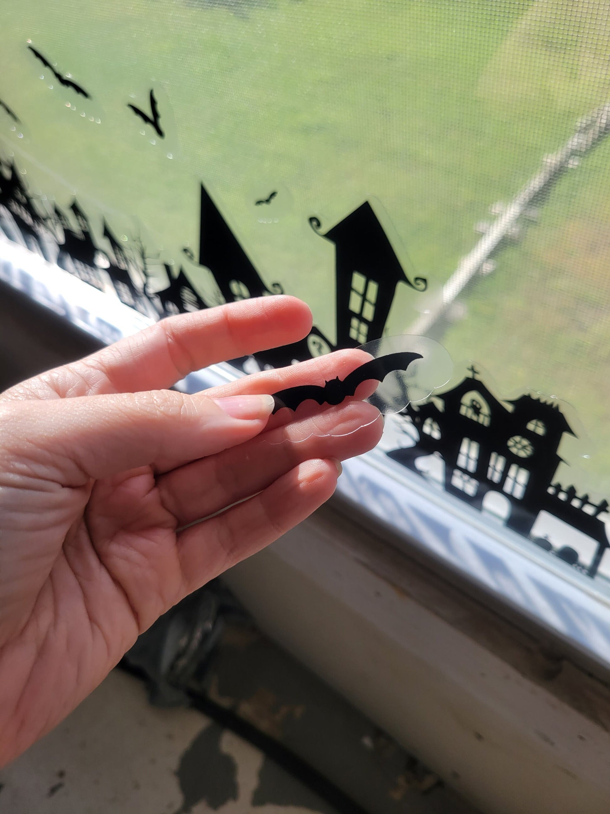 A bat from our Spooky Houses Static Window Cling Set, held in a hand with the rest of the set on a window in the background.