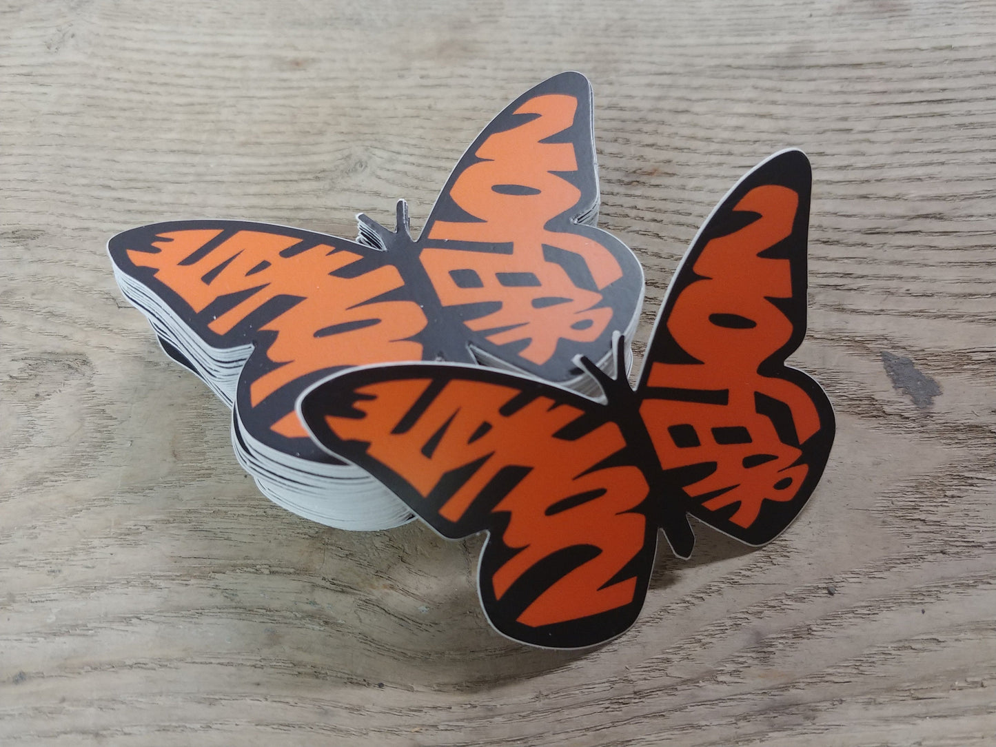 A stack of decals in the shape of a monarch butterfly. The wings, instead of the usual shapes of a Monarch, spell out the words "No Hate" and "No Fear" in orange. The text is bold and stands out against the design, emphasizing a message of peace and positivity. This is a pro-immigration decal.