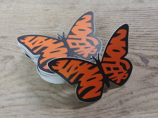 A stack of decals in the shape of a monarch butterfly. The wings, instead of the usual shapes of a Monarch, spell out the words "No Hate" and "No Fear" in orange. The text is bold and stands out against the design, emphasizing a message of peace and positivity. This is a pro-immigration decal.