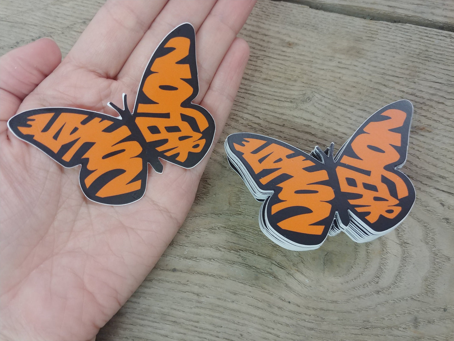Our No Hate No Fear Butterfly in a stack, and one held in hand for scale. This decal is in the shape of a monarch butterfly. The wings, instead of the usual shapes of a Monarch, spell out the words "No Hate" and "No Fear" in orange. The text is bold and stands out against the design, emphasizing a message of peace and positivity. This is a pro-immigration decal.
