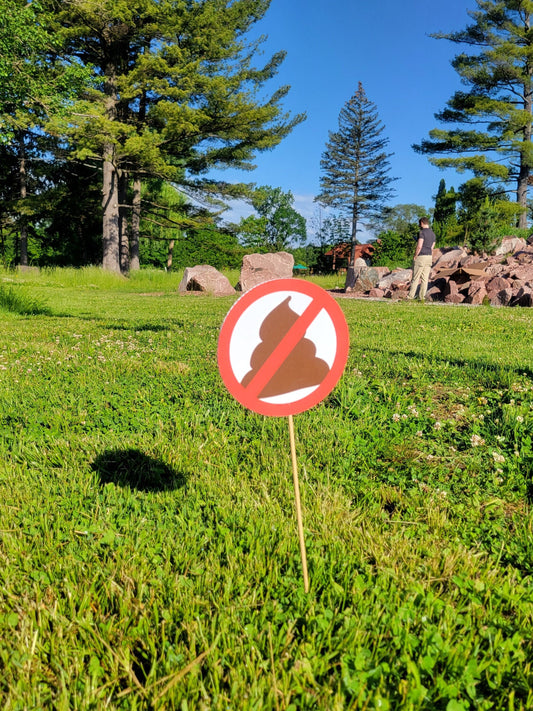 a photo of a no pooping yard sign (round, 6 inch diameter) on a wood dowel, in the grass of a public park.