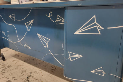 A blue steel desk with paper airplane decals applied all over, with solid lines behind them indicating their flight path. Cut from white vinyl.