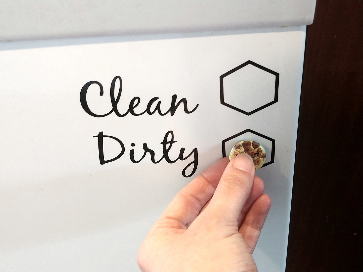 A hand is placing a magnet on the Dirty hexagon of an InkBird Print Studio LLC Clean/Dirty Dishwasher Vinyl Cut Decal, featuring the words Clean and Dirty alongside two hexagonal shapes. This decal uses permanent adhesive to function as an effective chore reminder for busy households.