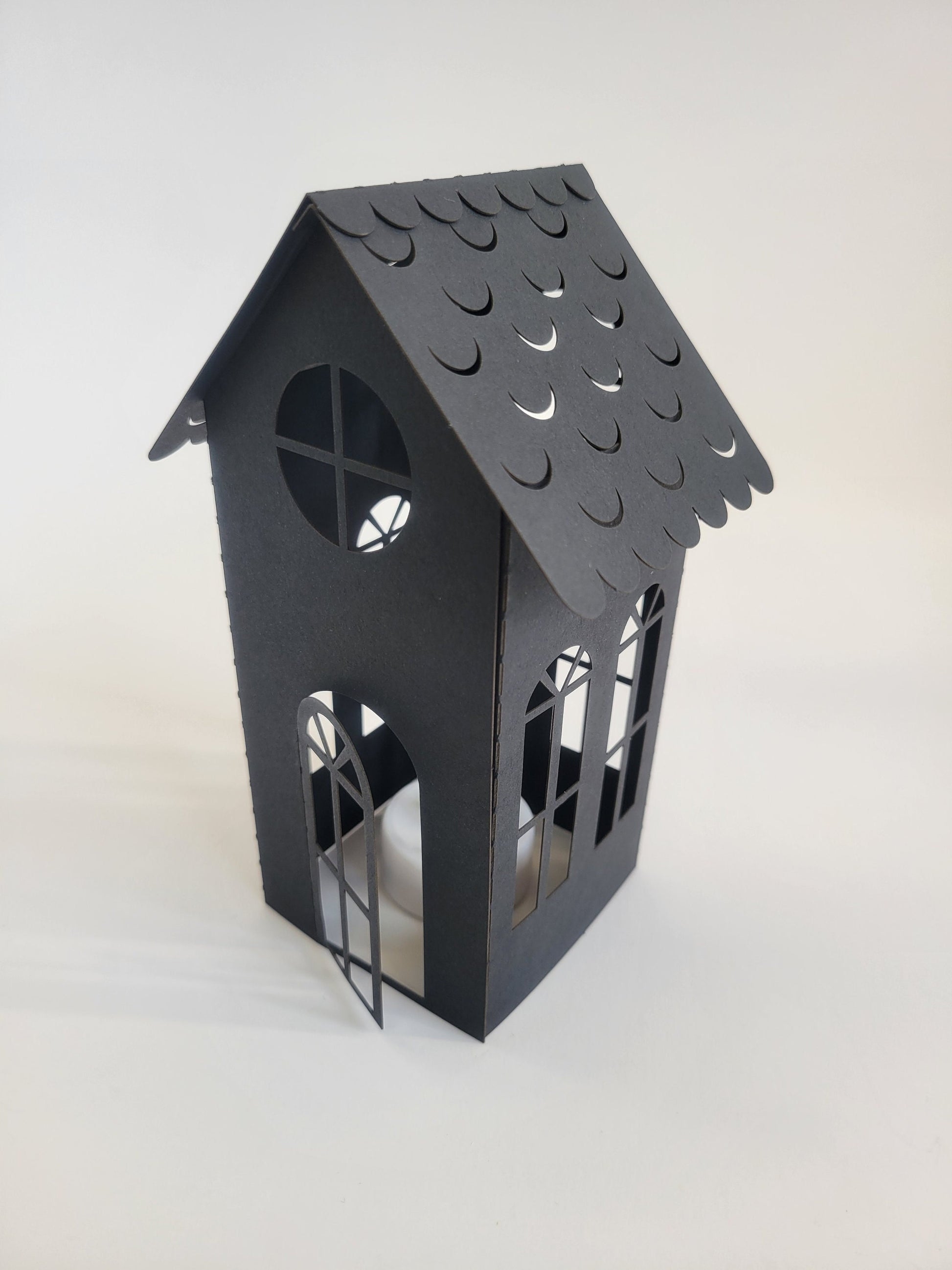 The DIY Spooky House Paper Kit by InkBird Print Studio LLC includes a small, black cardstock house with laser-cut decorative windows and a scalloped roof. An open door is designed to accommodate an LED tealight, creating an enchanting Halloween ambiance. The contrast between the white background and the kit's dark color highlights its striking appearance.