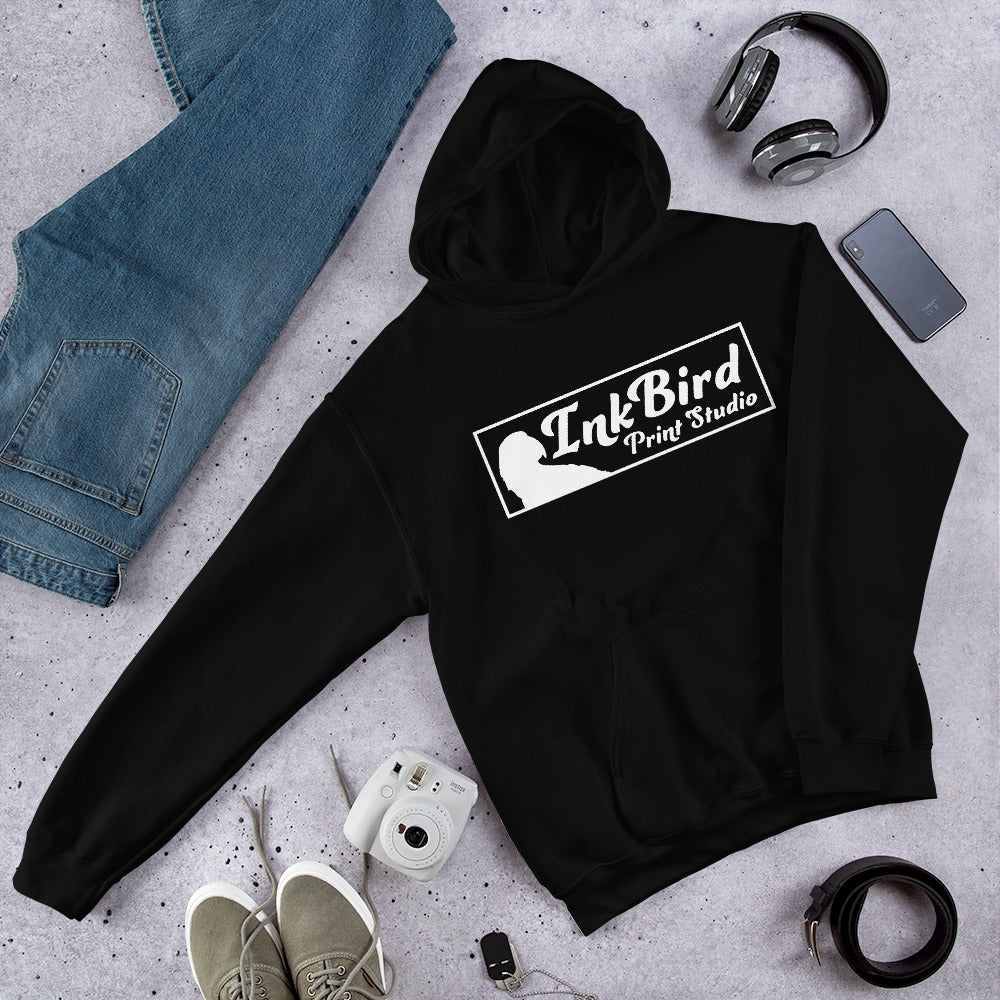 A stylish InkBird Print Studio Unisex Hoodie in black, featuring the brand InkBird Print Studio LLC printed in white, is laid out on a gray surface. Surrounding this cozy hoodie with a soft feel are blue jeans, green sneakers, a white instant camera, headphones, and a smartphone.